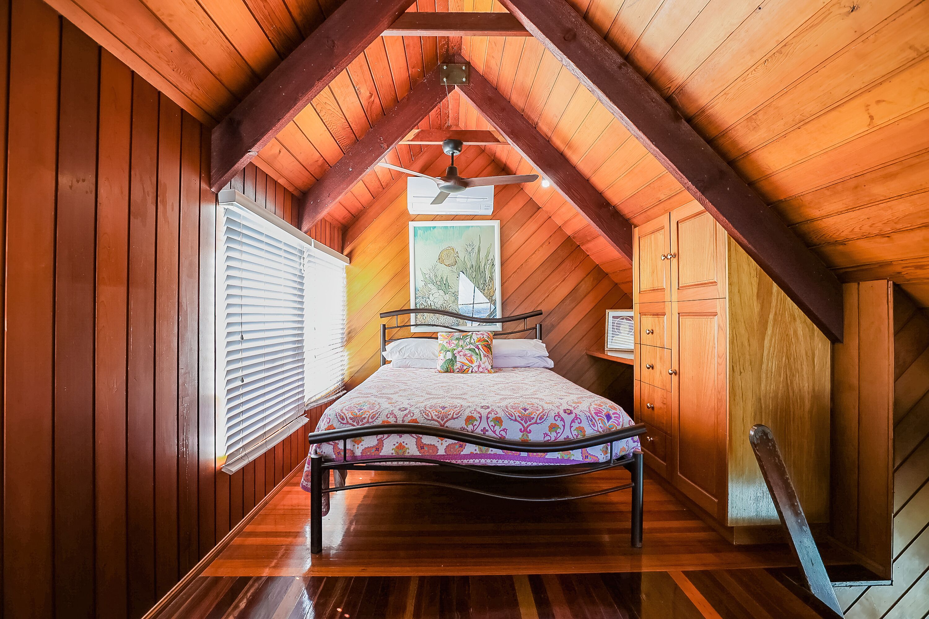 A bedroom with a sloped ceiling, wooden walls, a bed with a floral bedspread, a ceiling fan, and a window with blinds.