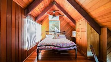 A bedroom with a sloped ceiling, wooden walls, a bed with a floral bedspread, a ceiling fan, and a window with blinds.