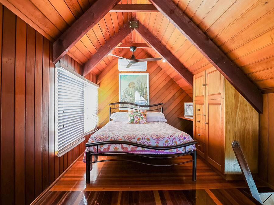 A bedroom with a sloped ceiling, wooden walls, a bed with a floral bedspread, a ceiling fan, and a window with blinds.