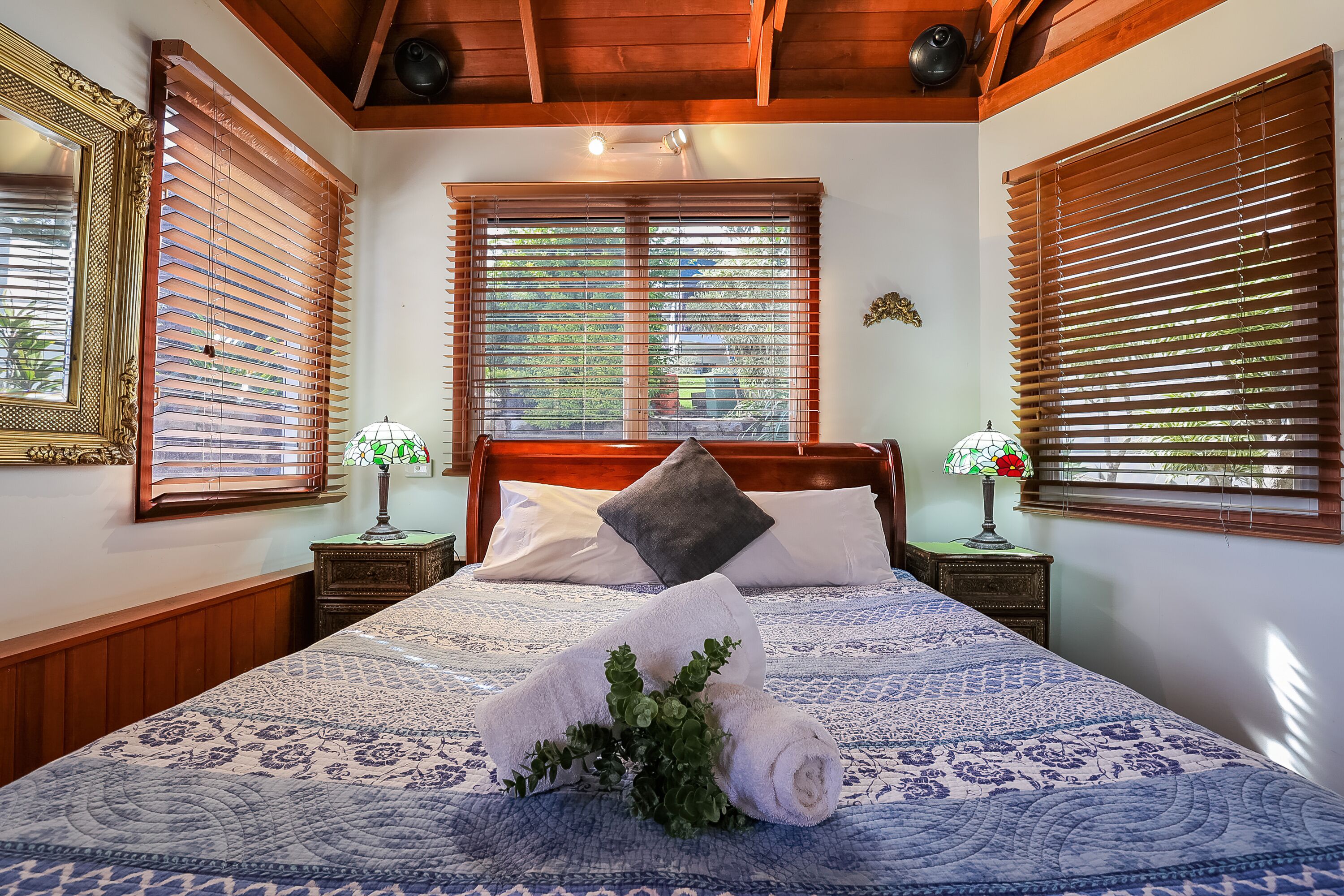 A neatly made bed with a patterned bedspread, a rolled towel, and a decorative plant. There are two bedside tables with lamps, a large mirror, and wooden blinds on the windows.