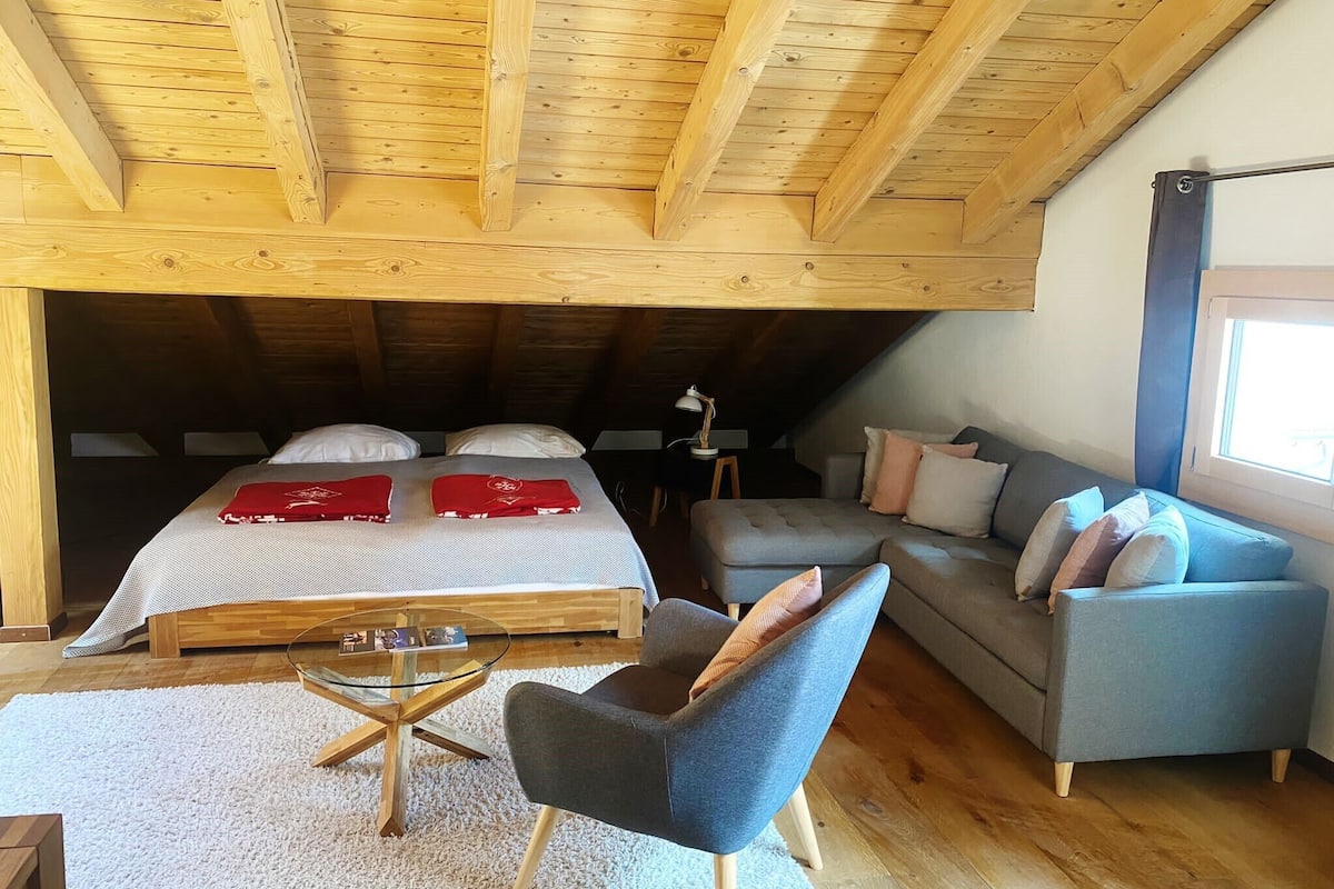 A cozy attic bedroom with a wooden ceiling, a large bed with red bedding, a grey sofa, and a small round table.