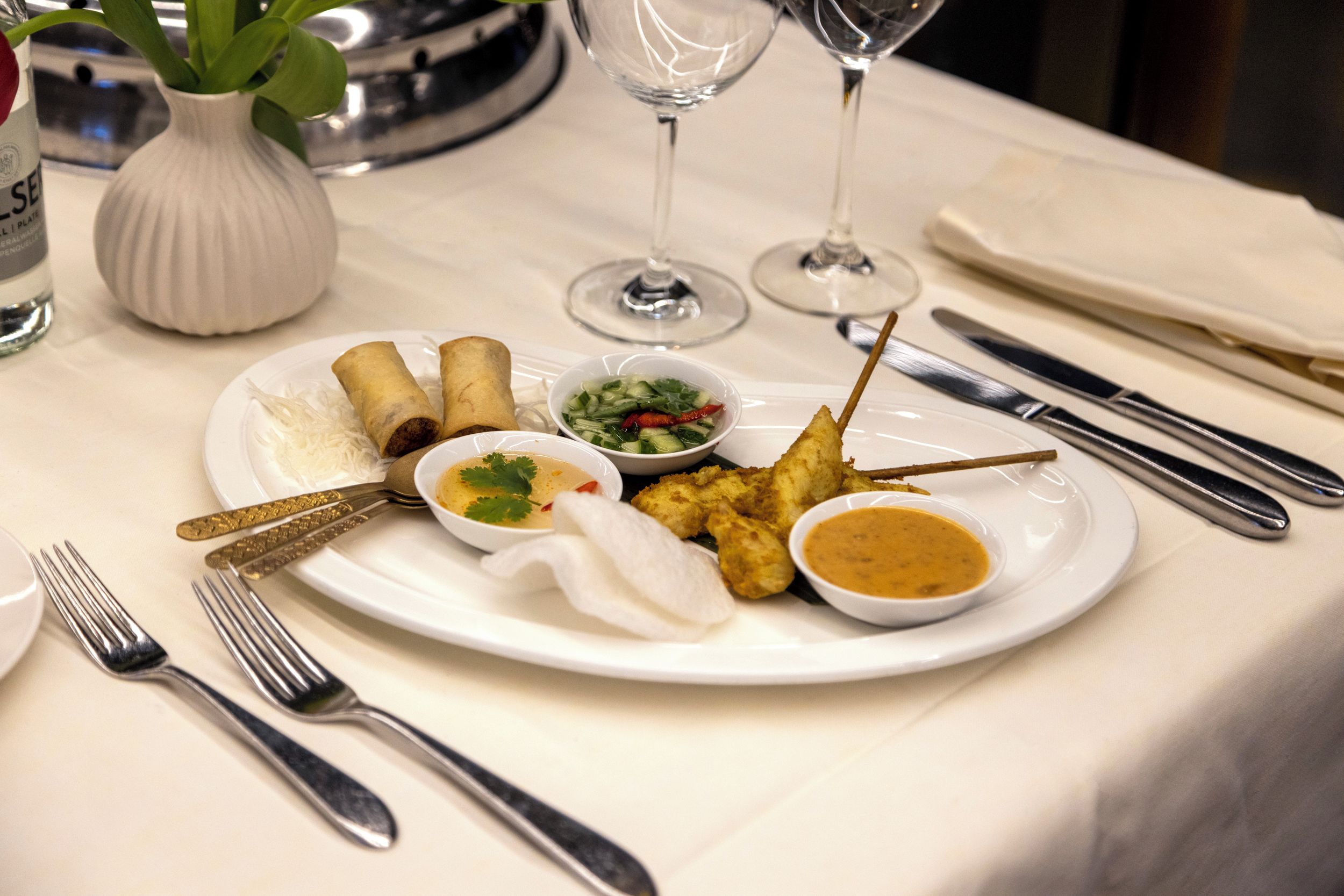 A dining table with a plate of food, including spring rolls, a soup, and a fish dish, accompanied by a small bowl of sauce and a side of vegetables.
