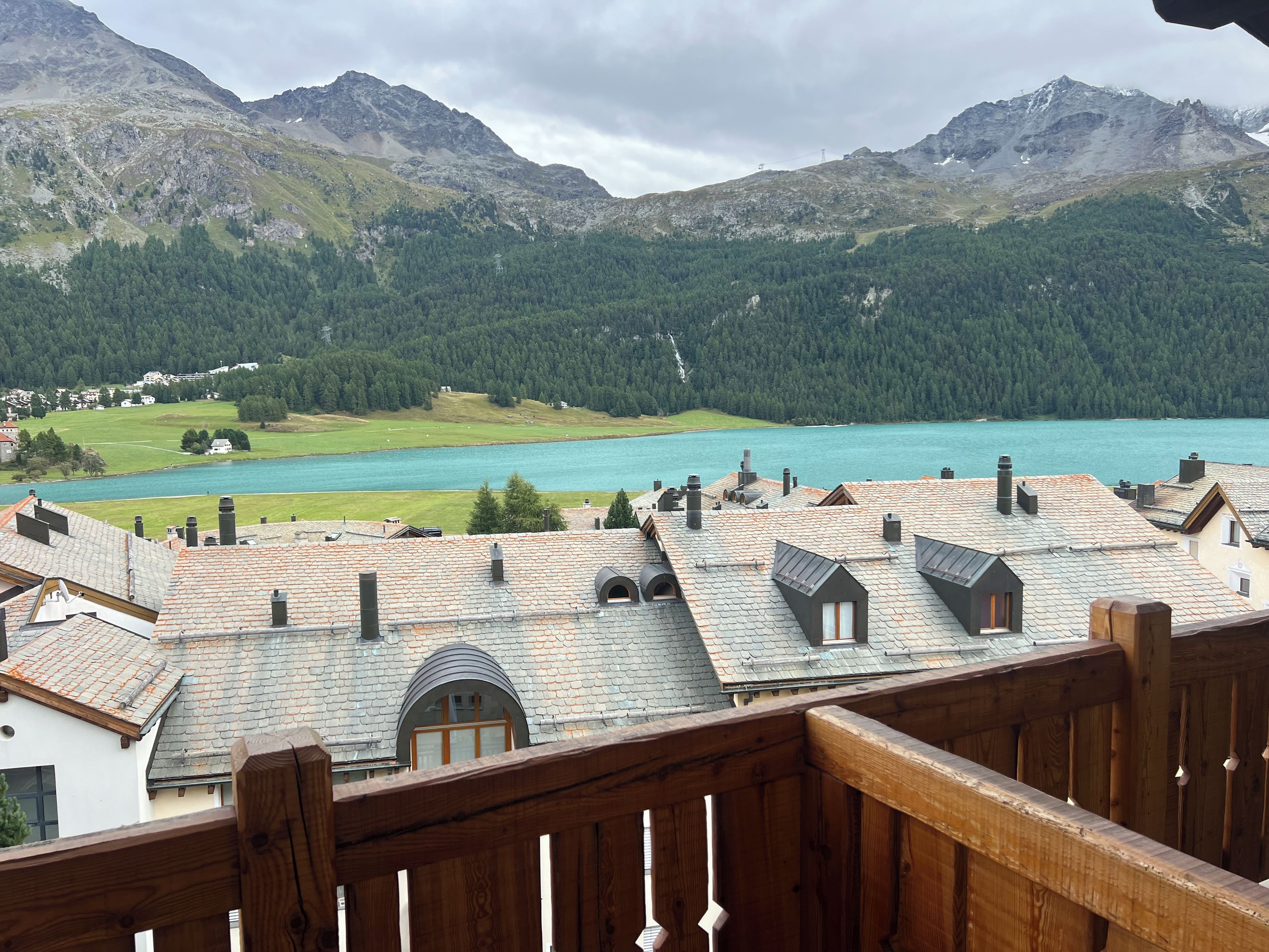 A mountainous landscape with a lake, a view of a residential area with houses, and a wooden fence in the foreground.