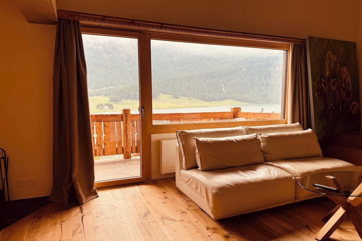A room with a wooden floor, a beige sofa, a painting on the wall, and a view of a lake and mountains through a large window.