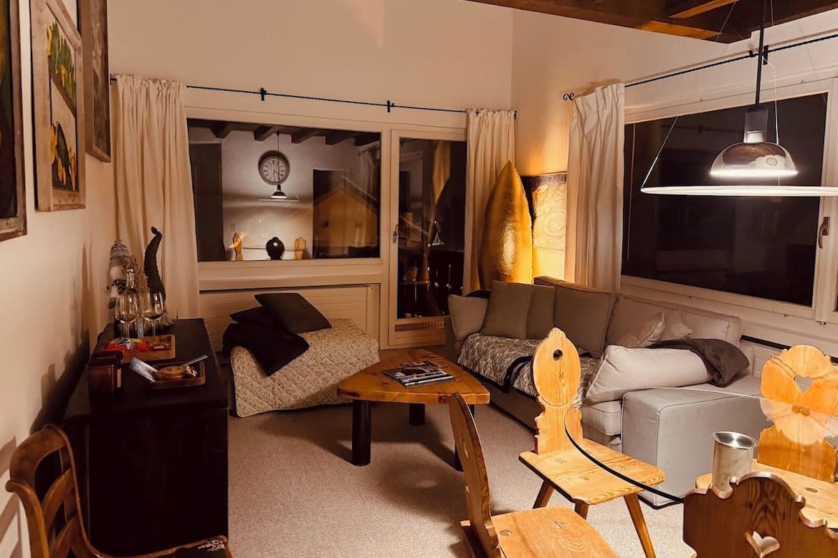 A cozy living room with a sofa, a wooden coffee table, and a dining table with chairs. There is a window with curtains, a lamp, and a clock on the wall.