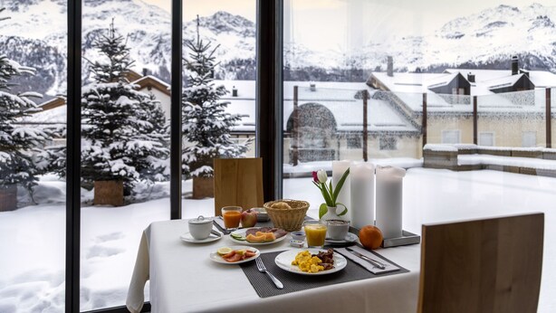 Een ontbijttafel met diverse etenswaren en zicht op een besneeuwd berglandschap door grote ramen.