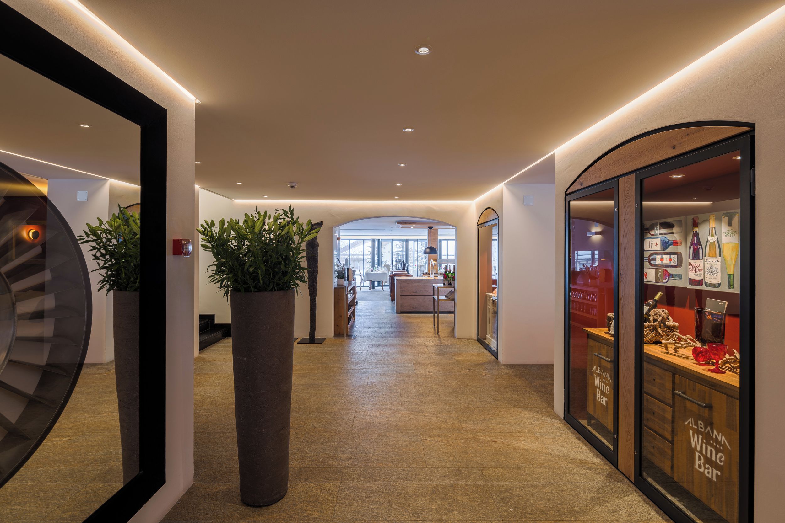 A hotel corridor with a bar on the right, a staircase on the left, and a large plant in the center.