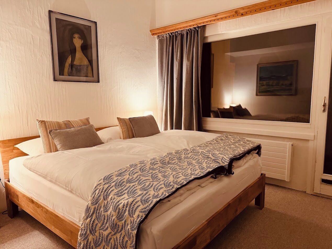 A neatly made bed with a patterned throw, a wooden headboard, and a large window with curtains.