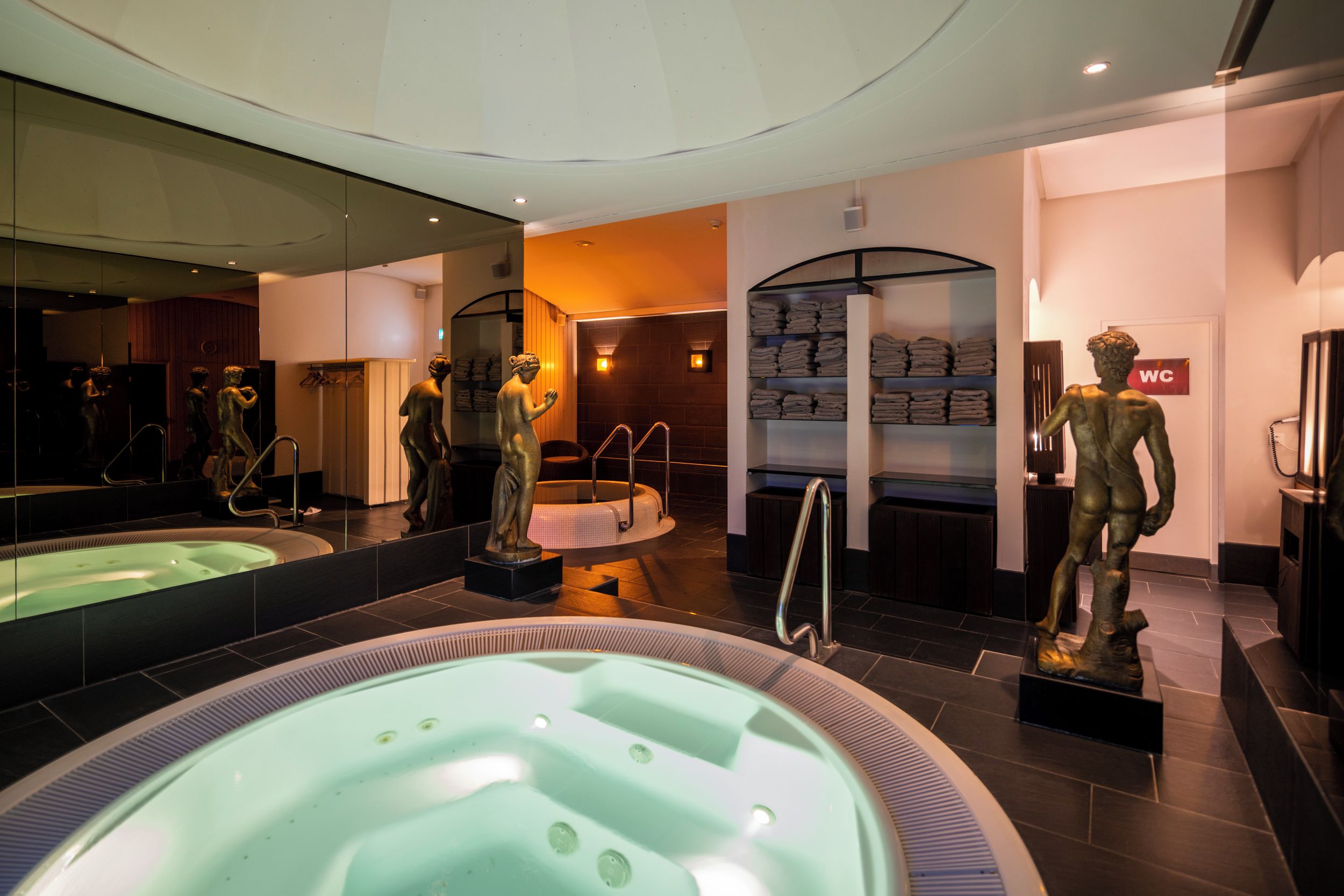 A modern bathroom with a large mirror, a hot tub, and a statue.