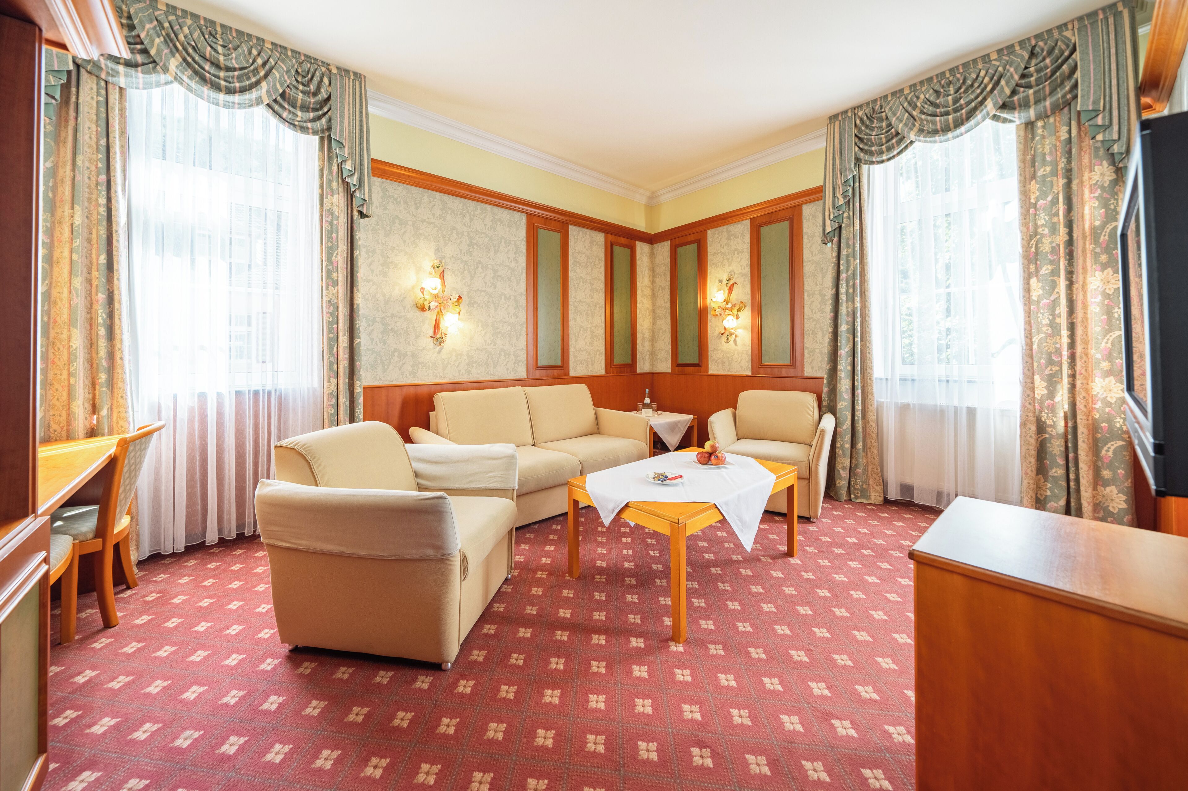 Hotelkamer met een zithoek, een bijzettafeltje, een bureau en een kamerbreed tapijt met dessin.