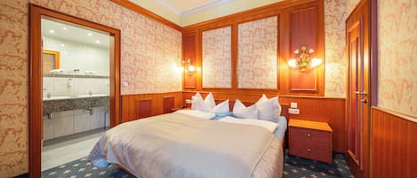A hotel room with a large bed, wooden headboard, two bedside tables, a nightstand, a mirror, and a bathroom visible through an open door.