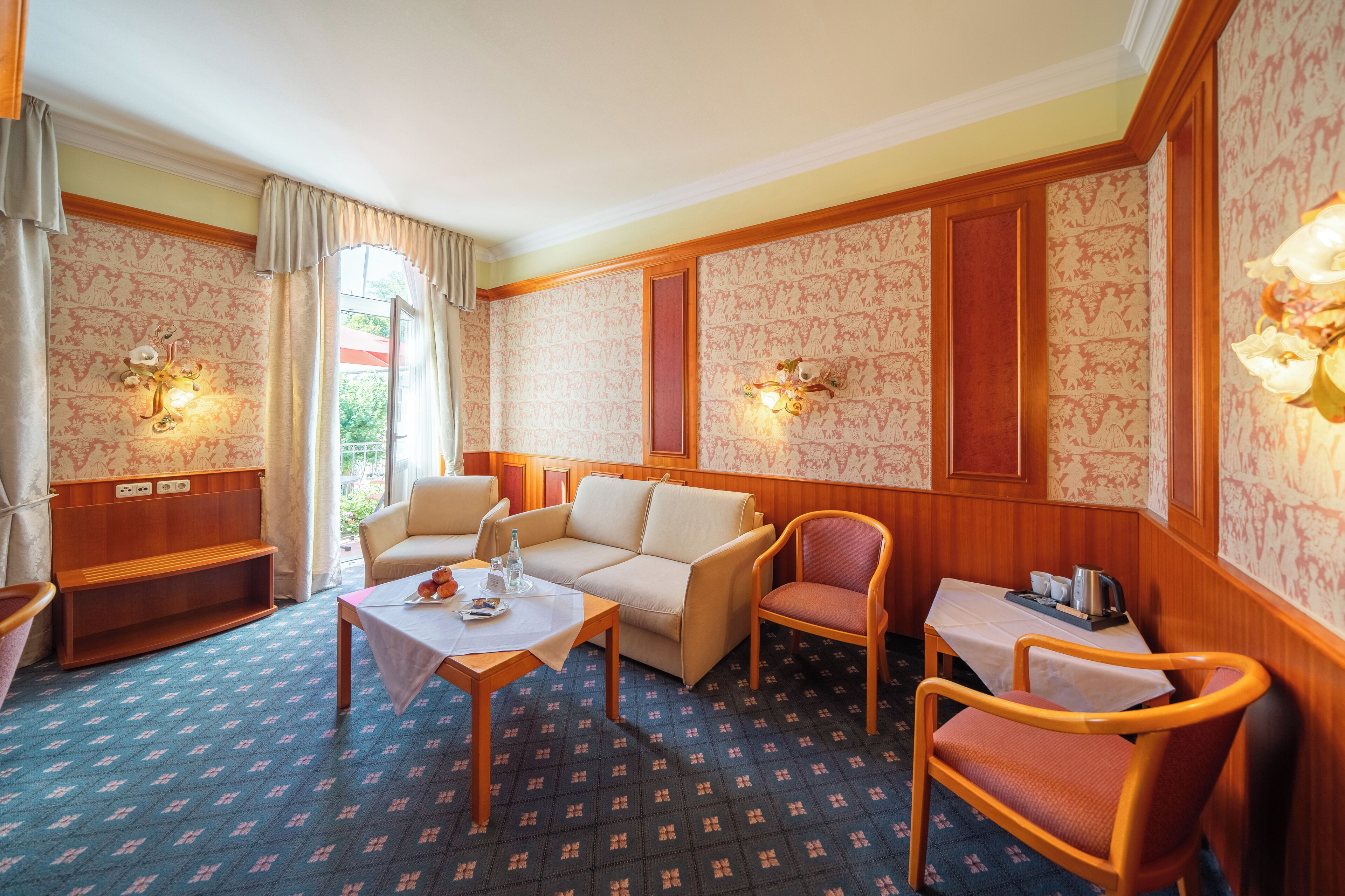 Une chambre d’hôtel avec un canapé, des fauteuils, une petite table, un banc et un papier peint à motifs.