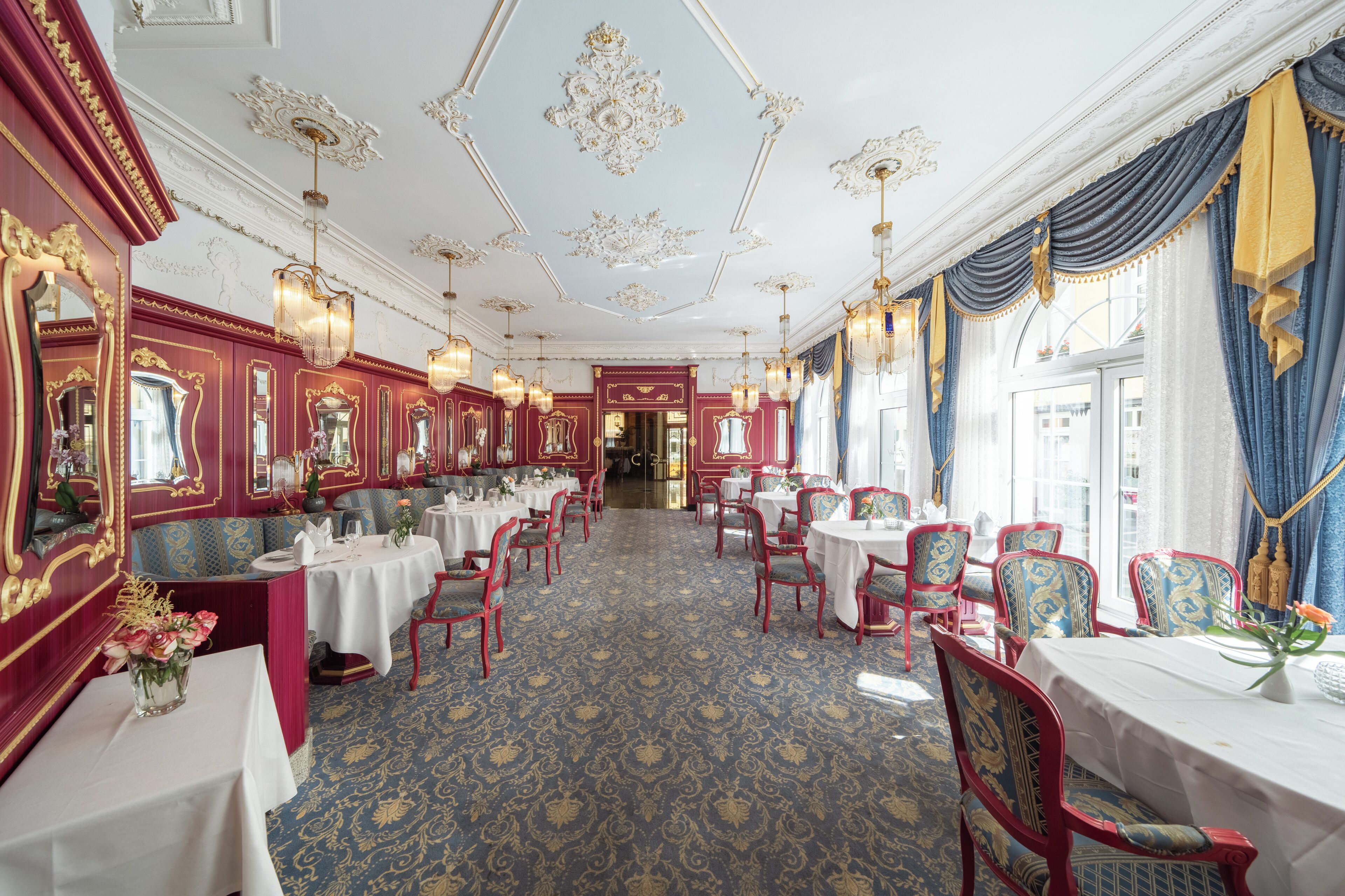A dining area with tables and chairs, ornate ceiling decorations, and a patterned carpet.