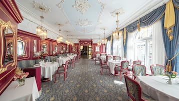 A dining area with tables and chairs, ornate ceiling decorations, and a patterned carpet.
