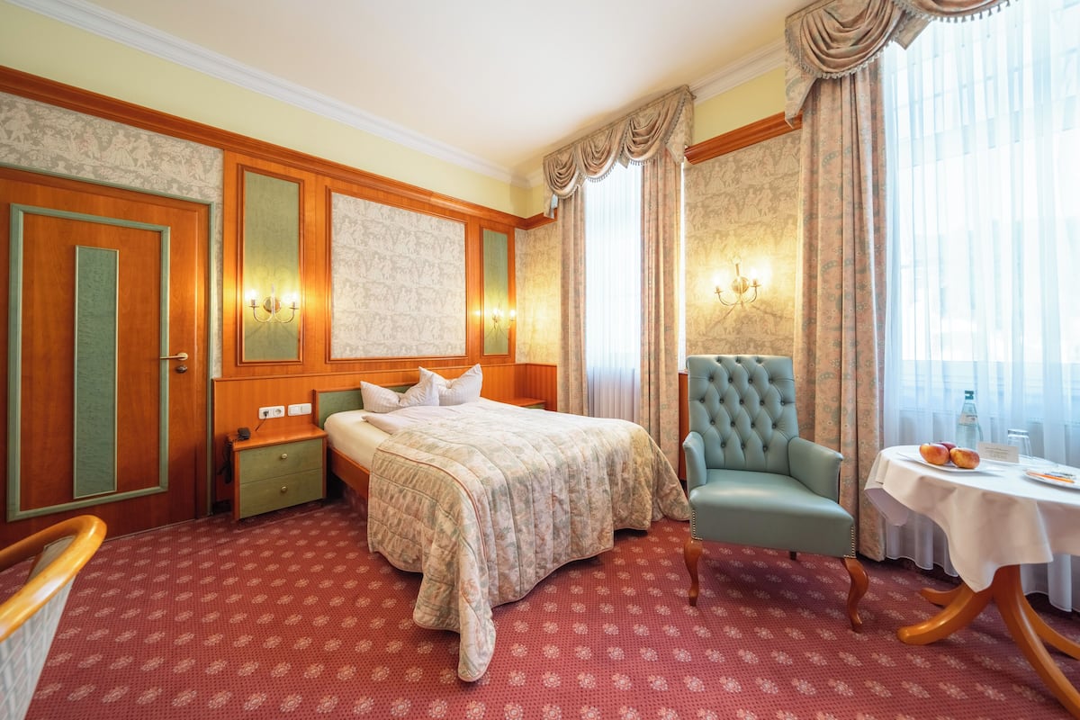 Ein Hotelzimmer mit Bett, einem Sessel, einem Tisch mit Flasche und Gläsern sowie einem Fenster mit Vorhängen.