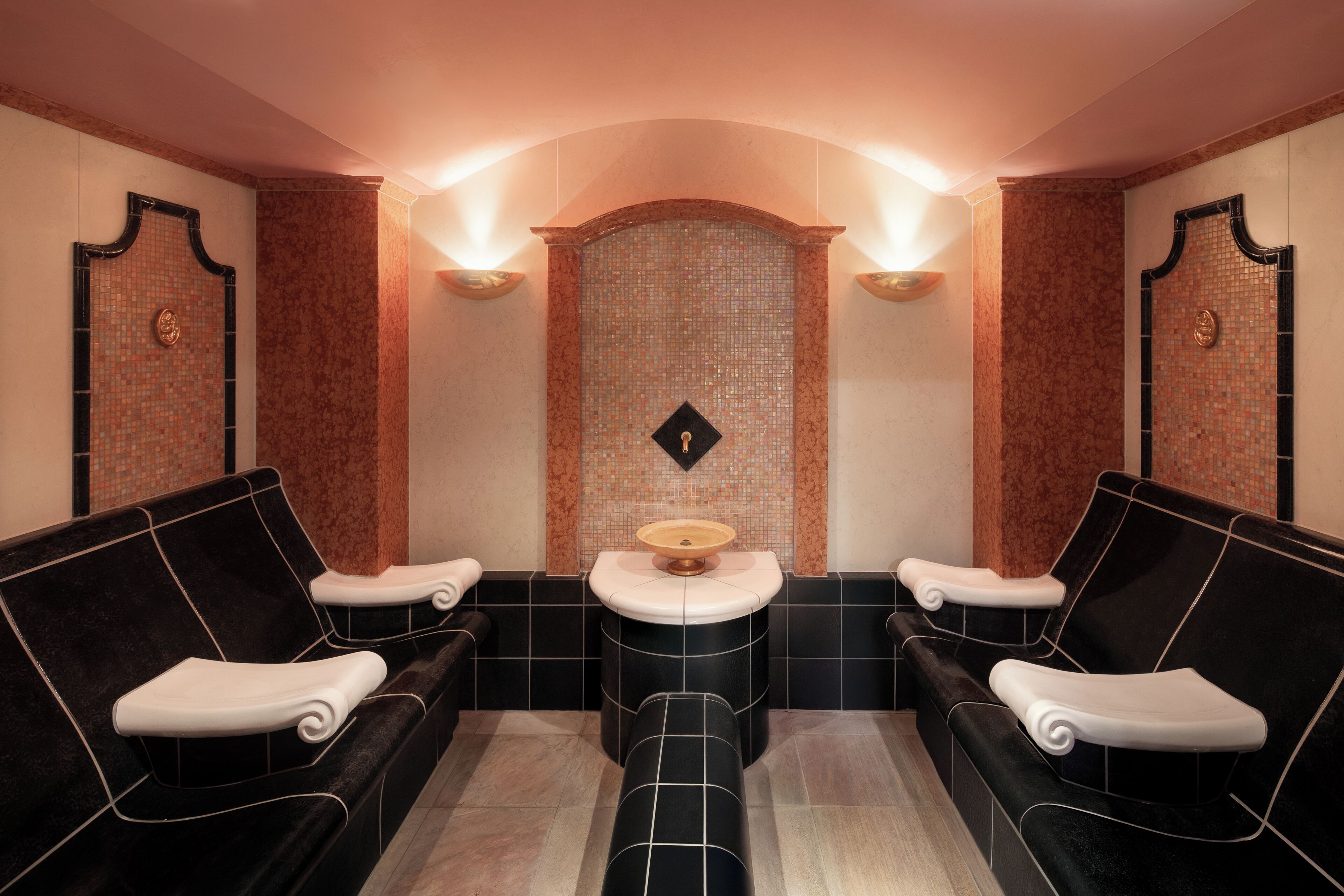 A spa room with two black sofas, a central sink, and a mosaic wall.