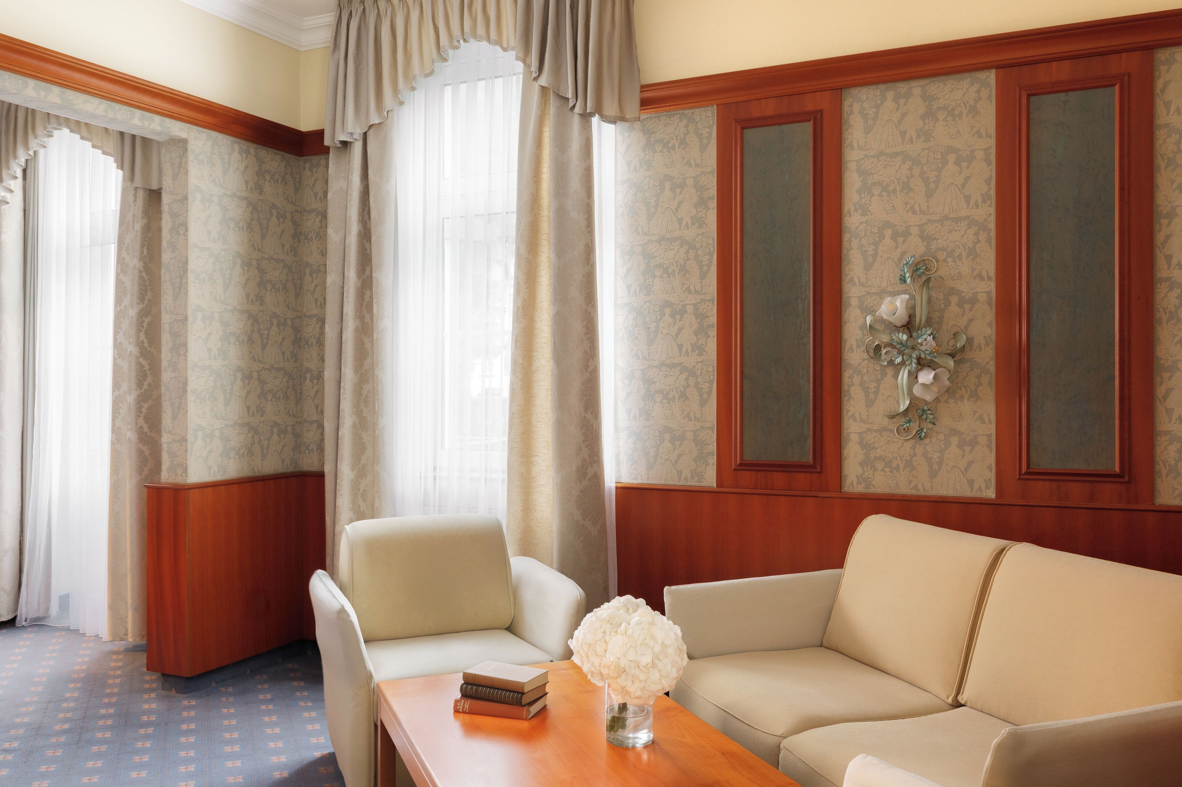Ein Hotelzimmer mit einem beige farbenen Sofa, einem Holztisch mit einem Buch und einer Vase sowie einem großen Fenster mit Vorhängen.