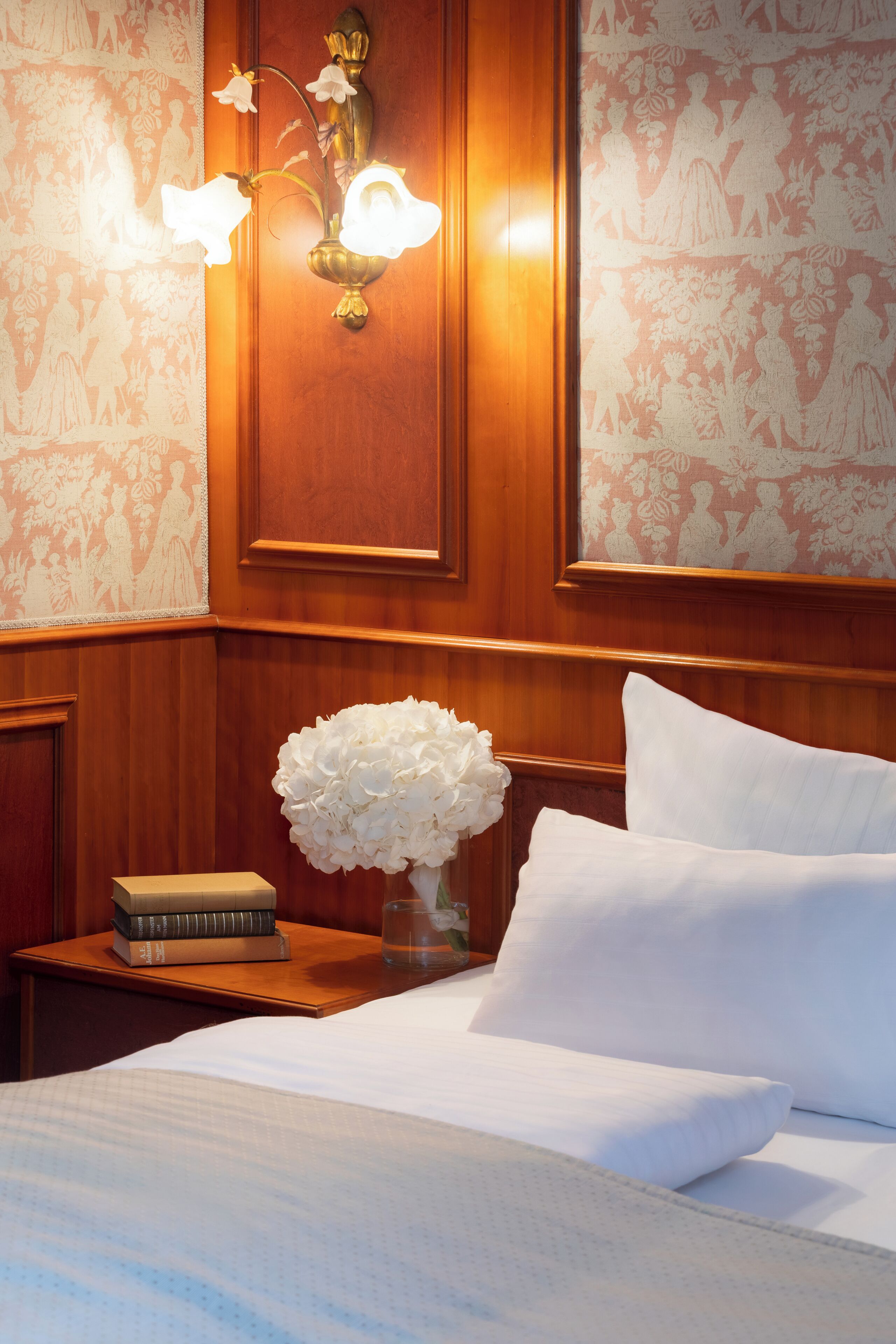 A hotel room with a wooden headboard, a bedside table with a vase of flowers, a stack of books, and a neatly made bed.