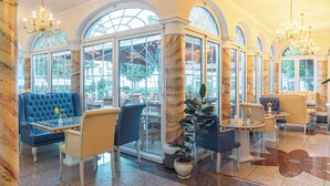 A luxurious lobby with marble columns, blue upholstered seating, and large windows offering views of the outdoors. - Hotel Fürstenhof Bad Bertrich (Bad Bertrich)