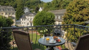 A balcony with wicker chairs, a table with breakfast, and a view of a hill with buildings. - Hotel Fürstenhof Bad Bertrich (Bad Bertrich)