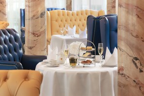 Een eettafel met een witte tafelkleed, een theeservies, een taartplateau en een glas wijn.