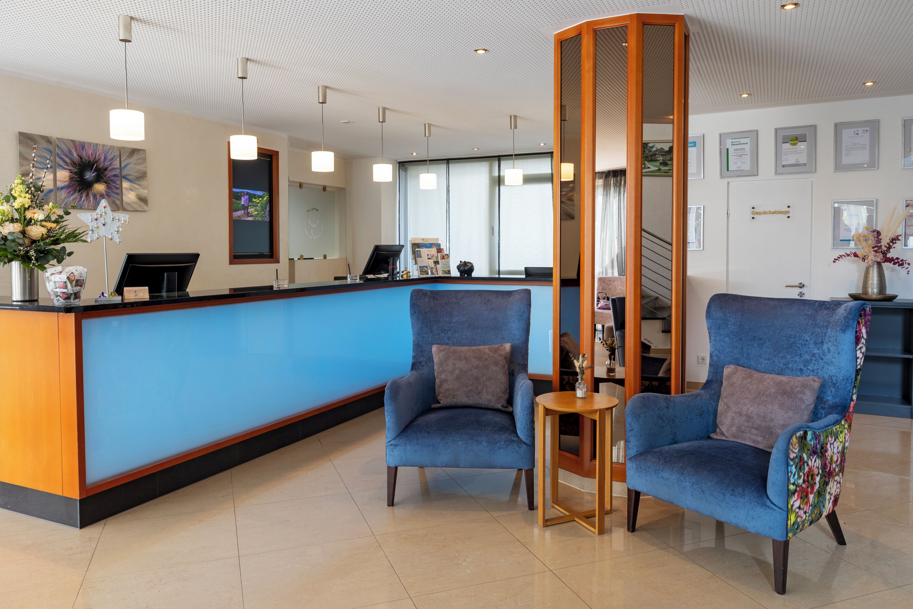 Un área de recepción con un mostrador azul, dos sillones azules, una mesita de madera y un jarrón decorativo.