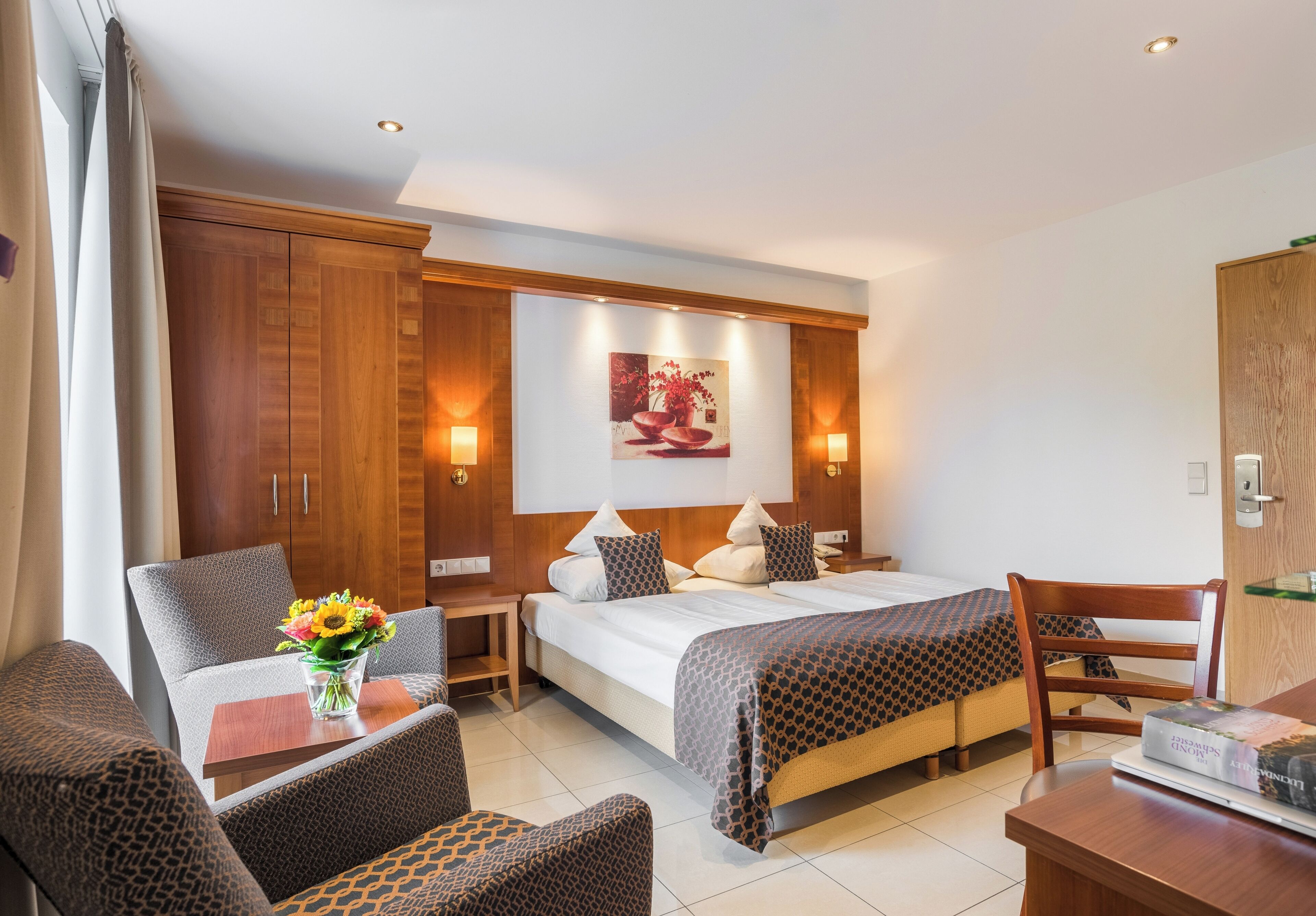 A hotel room with a bed, two armchairs, a small table with a vase of flowers, and a wooden wardrobe.