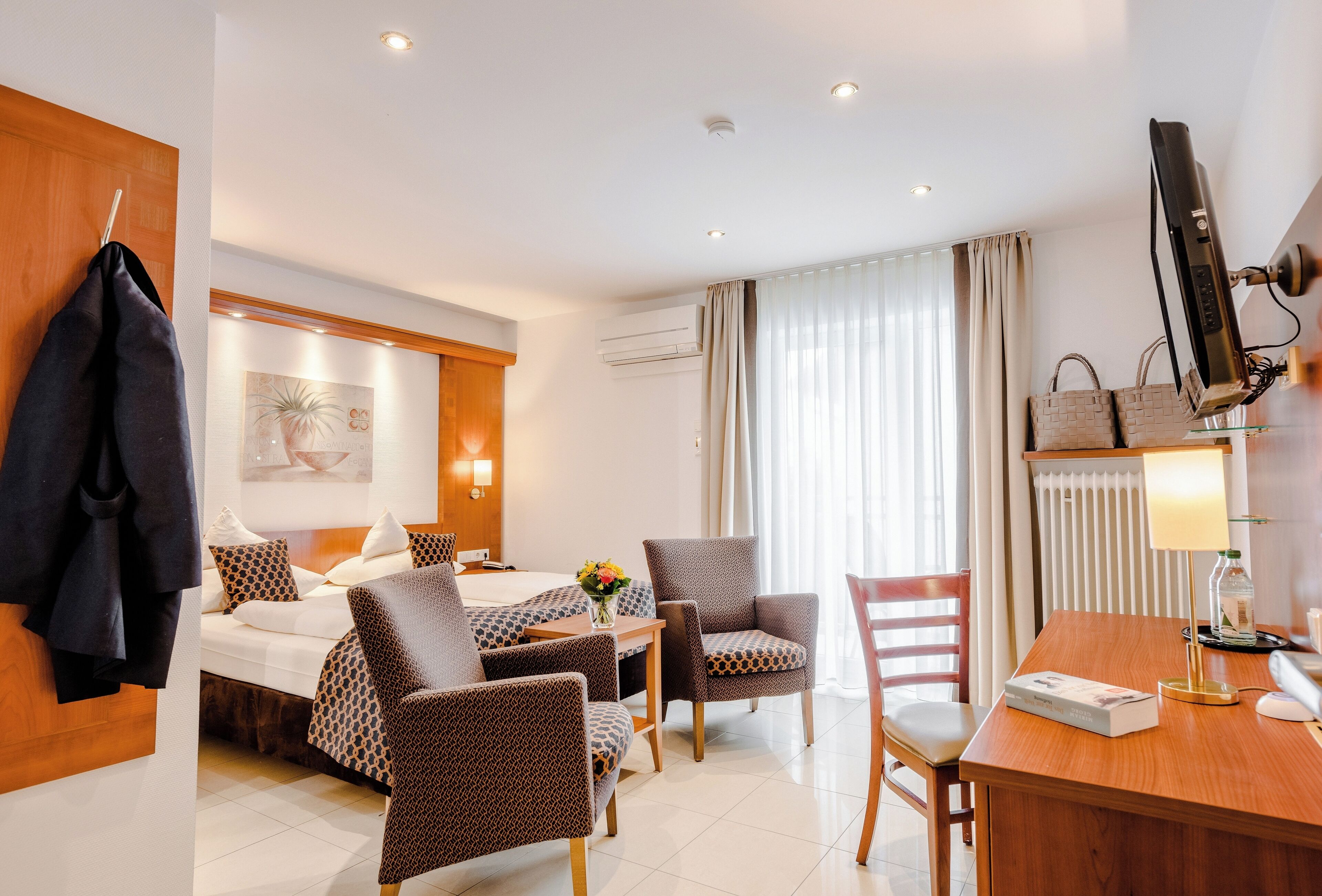 Ein Hotelzimmer mit Bett, zwei Sesseln, einem Schreibtisch, einem Fernseher und einem fensterbepflanzten Fenster.
