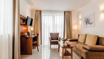 A hotel room with a flat-screen TV mounted on a wooden desk, a comfortable armchair, a brown sofa, and a small table with a vase of flowers.