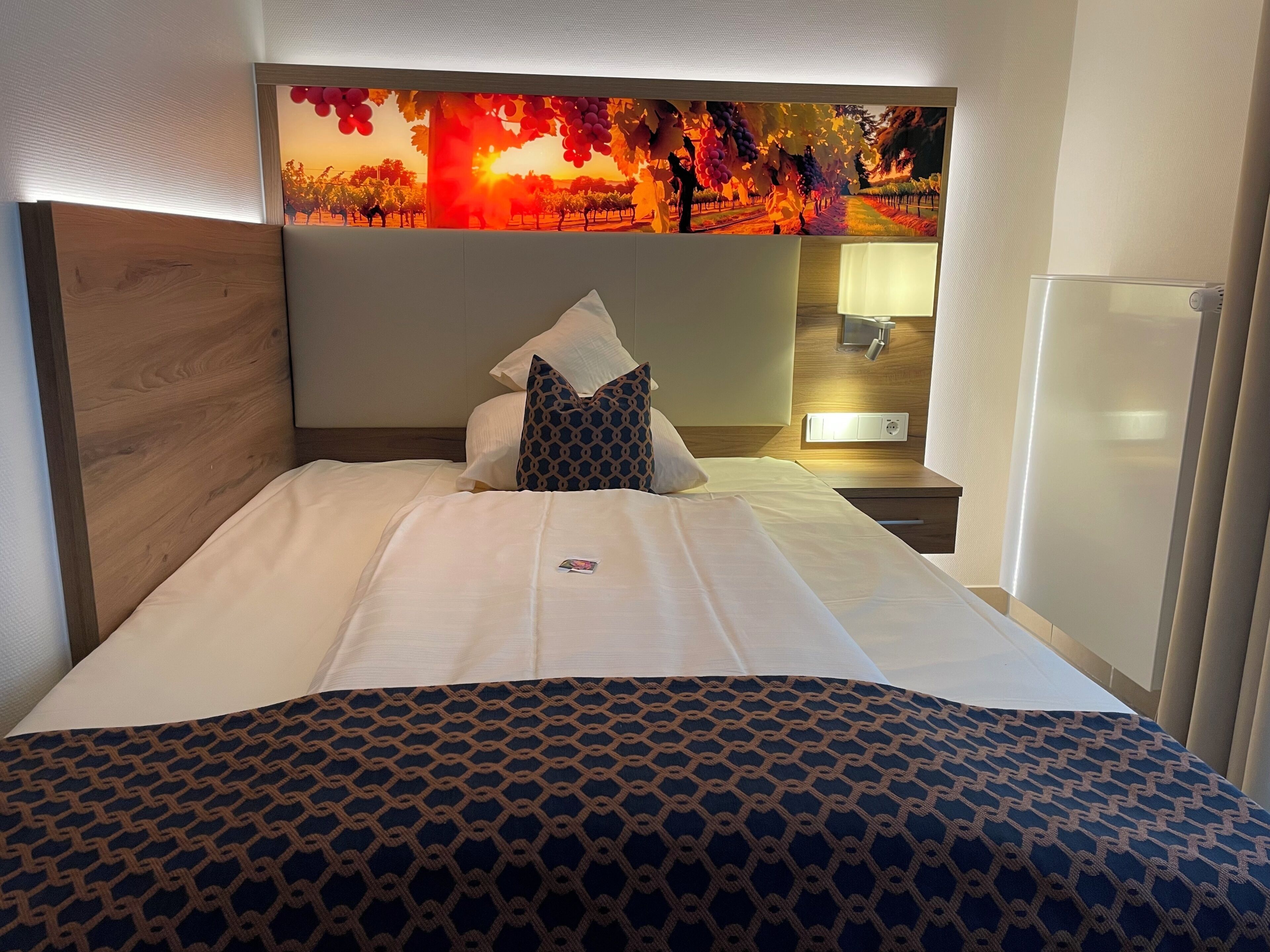 Una habitación de hotel con cama, cabecera con mural, mesita de noche y tocador.