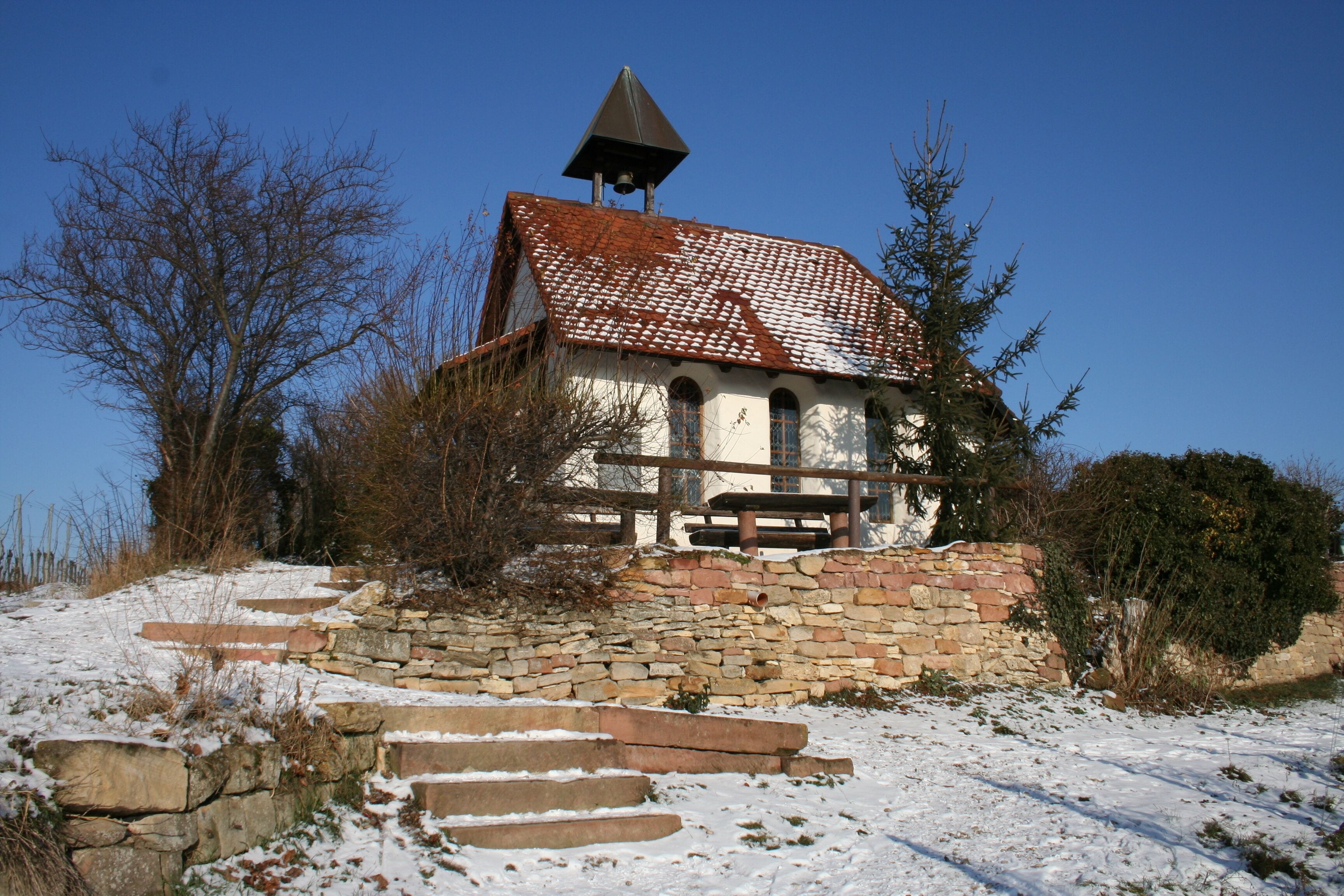 Una pequeña capilla de piedra con tejado de tejas y un campanario, rodeada de terreno y árboles cubiertos de nieve.