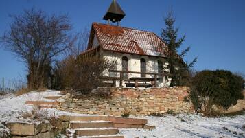 Una pequeña capilla de piedra con tejado de tejas y un campanario, rodeada de terreno y árboles cubiertos de nieve.