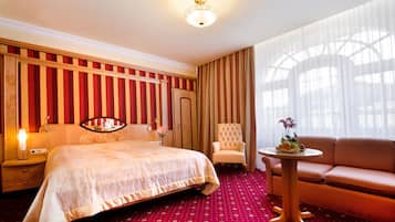 A hotel room with a large bed, a sofa, a chair, a small table with flowers, and a large window with curtains.