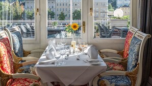 A dining table set with white tablecloth, glassware, and a sunflower centerpiece. The room has ornate chairs and a view of buildings through large windows. - Häcker's Grand Hotel Bad Ems (Bad Ems)