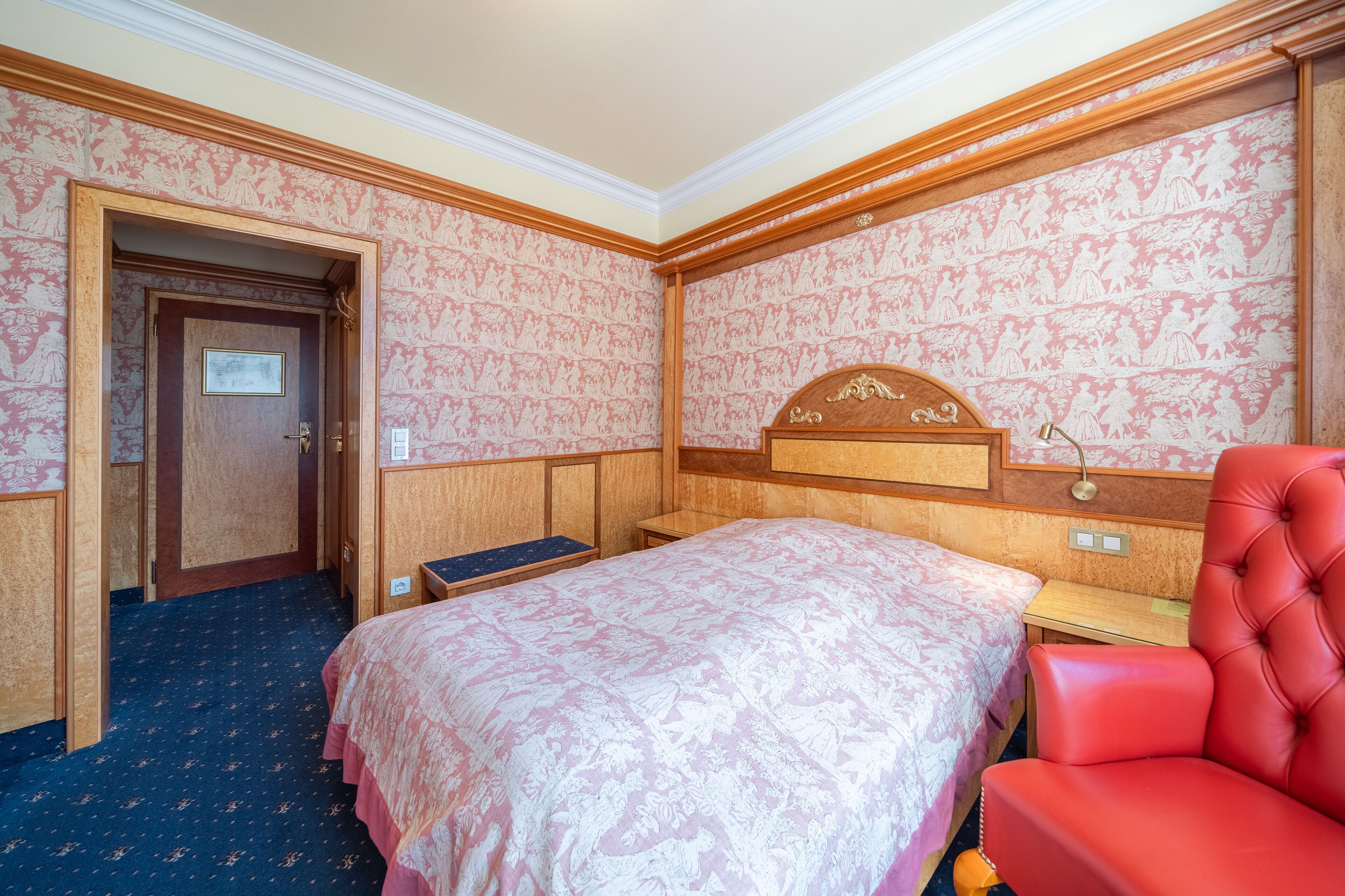 A hotel room with a large bed, a red armchair, a wooden headboard, and a door leading to another room.