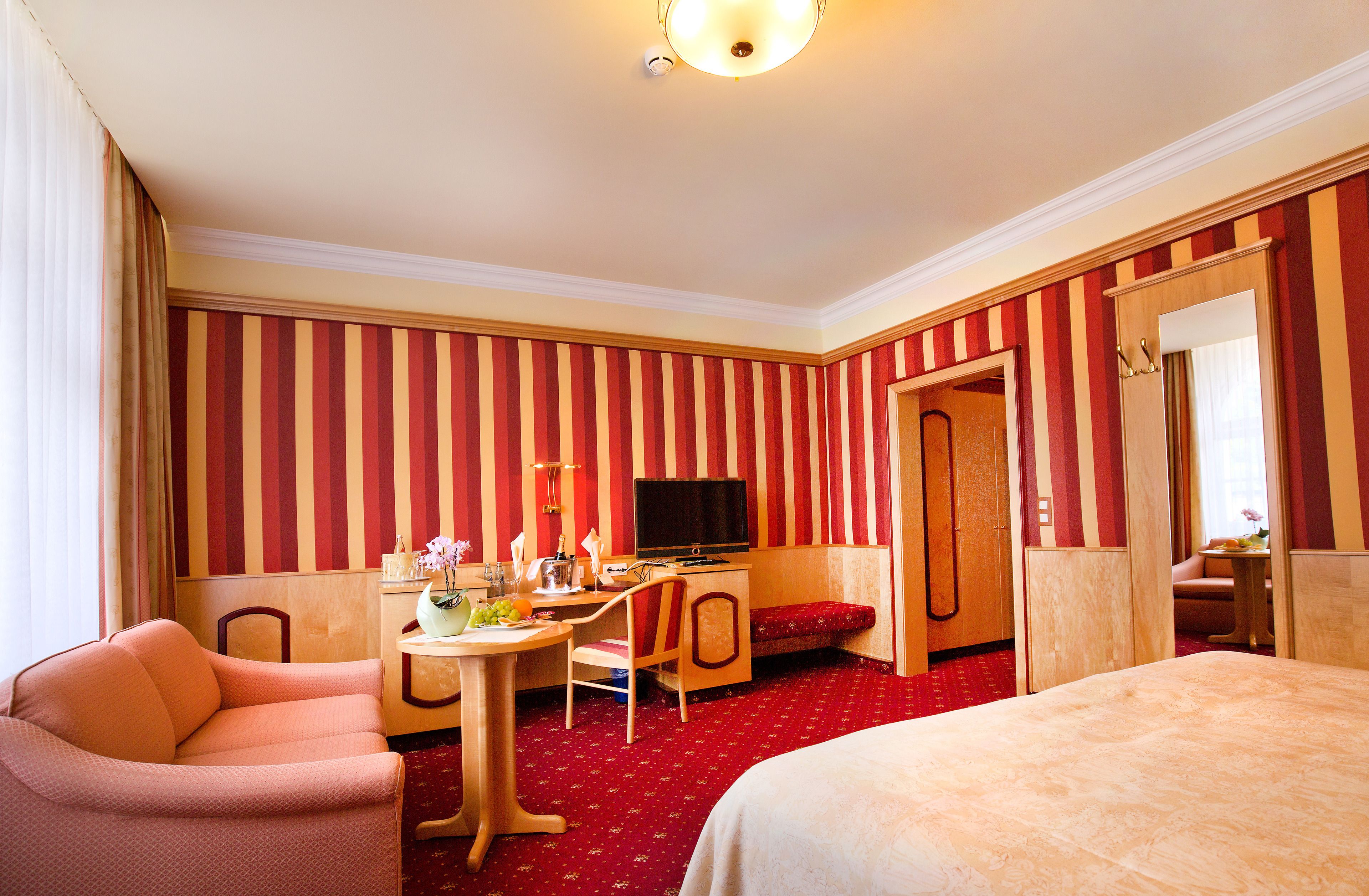 Ein Hotelzimmer mit gestreifter Tapete, einem Bett, einem Schreibtisch mit Stuhl, einem Fernseher und einem kleinen Tisch mit Blumen in einer Vase.