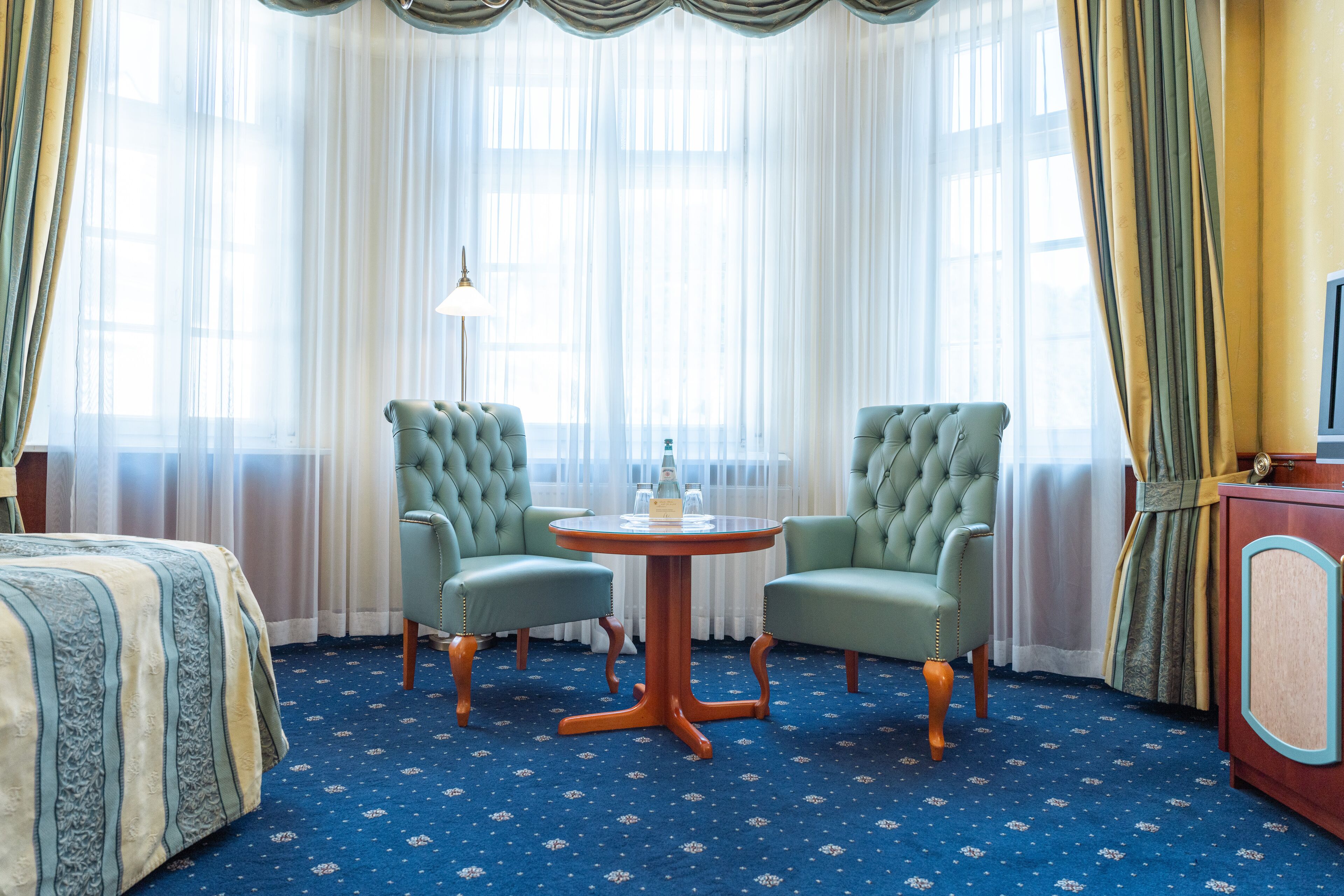 Habitación de hotel con una cama, dos sillones verdes, una mesa redonda, una lámpara y un televisor sobre un mueble.