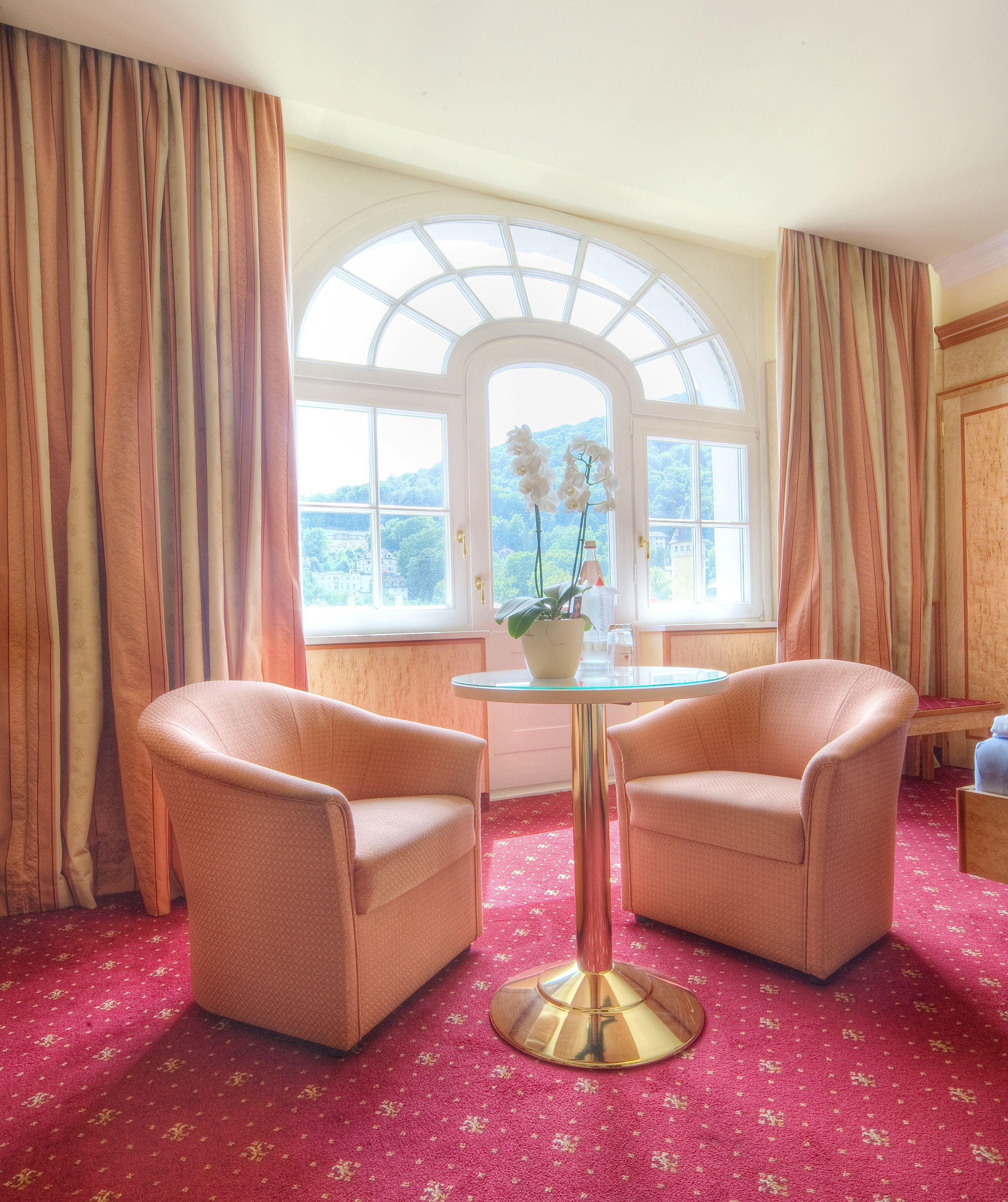 Ein Zimmer mit zwei Sesseln, einem Glastisch und Blick auf eine grüne Landschaft durch ein großes, rundbogenförmiges Fenster.