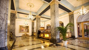 A lobby with marble columns, chandeliers, a reception desk, and a large painting. - Häcker's Grand Hotel Bad Ems (Bad Ems)