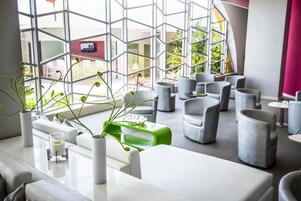 Un interior moderno con una zona de estar que incluye sofás y sillones grises, un mostrador de recepción blanco y un amplio ventanal con un diseño geométrico.