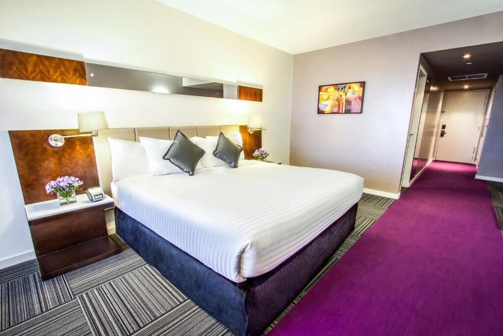 A hotel room with a large bed, a nightstand with a lamp, a small table with a vase of flowers, and a painting on the wall.