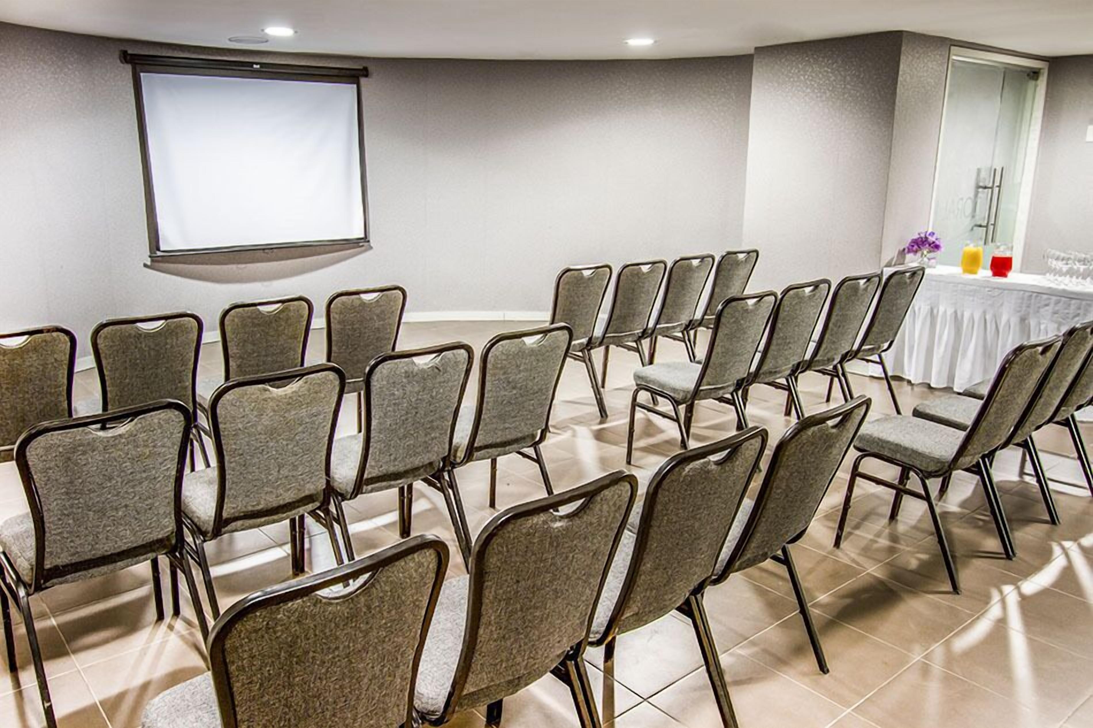 Un salón de conferencias con filas de sillas, una pantalla de proyector y una mesa con bebidas y un jarrón de flores.