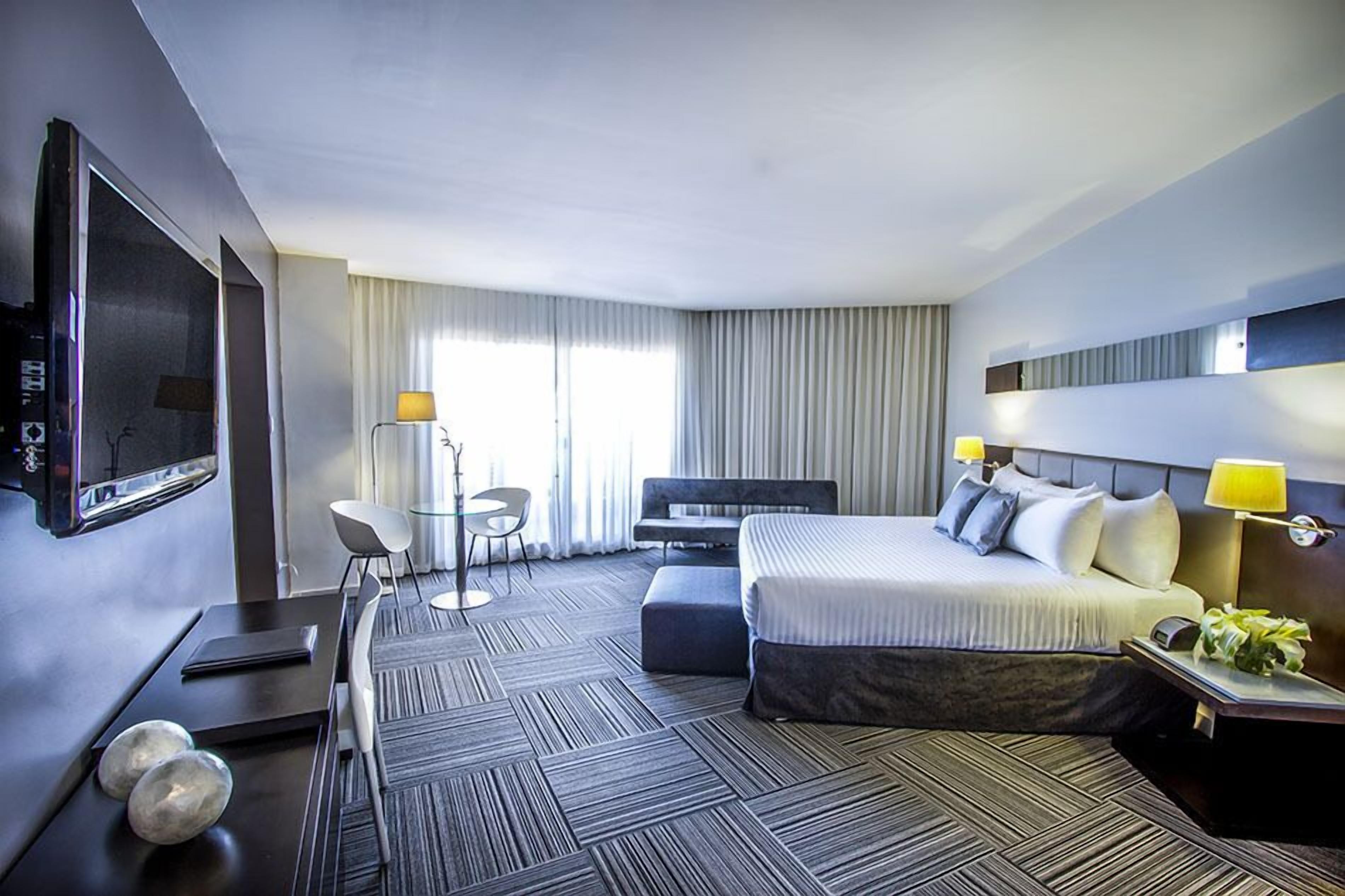 A modern hotel room with a large bed, a desk, a TV, and a seating area with a chair and a small table.