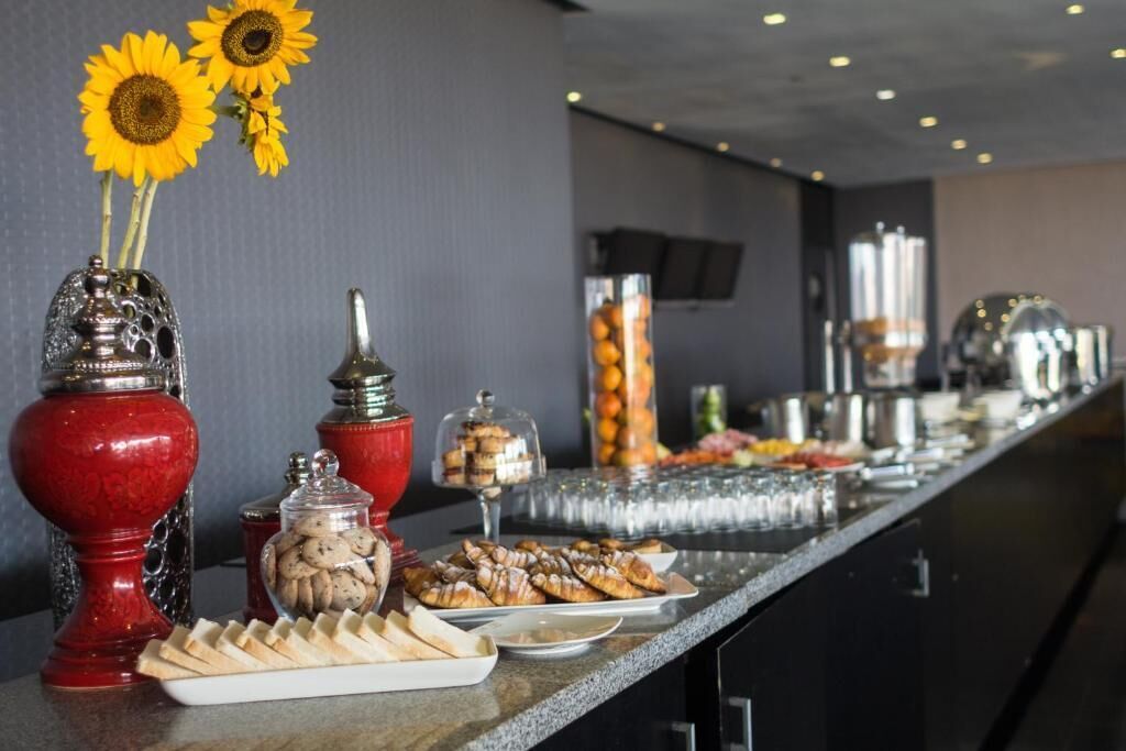 Montaje de bufé con diversas opciones de comida, incluyendo pasteles y un frasco de galletas, en un ambiente moderno de hotel.