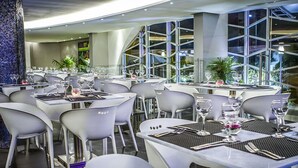 Un restaurante moderno con sillas y mesas blancas, ventanas grandes y un pilar decorativo.