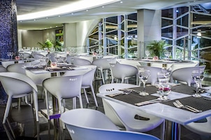 Un restaurante moderno con sillas y mesas blancas, ventanas grandes y un pilar decorativo.