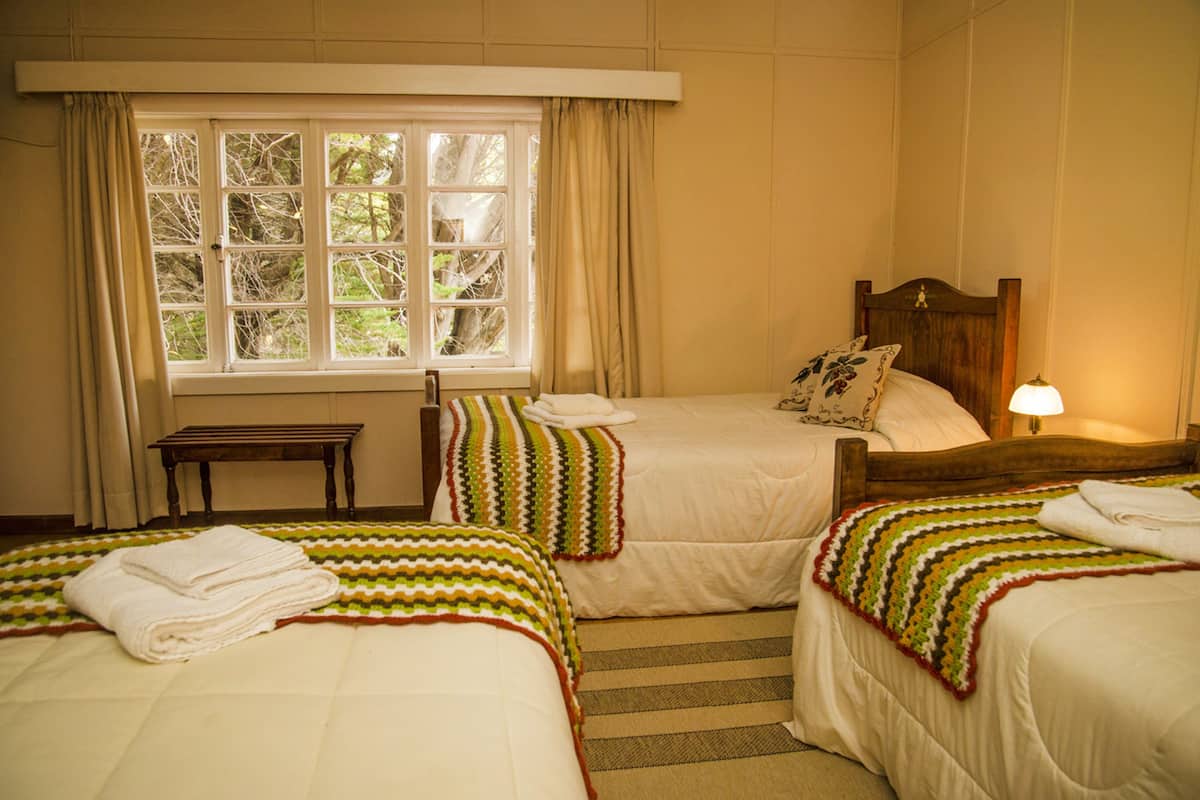 A hotel room with two beds, a window with curtains, a bedside table, and a lamp.