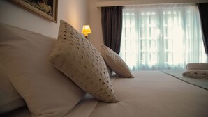 Een net opgemaakt bed met twee gestreepte kussens, een lamp op een nachtkastje en een raam met gordijnen.
