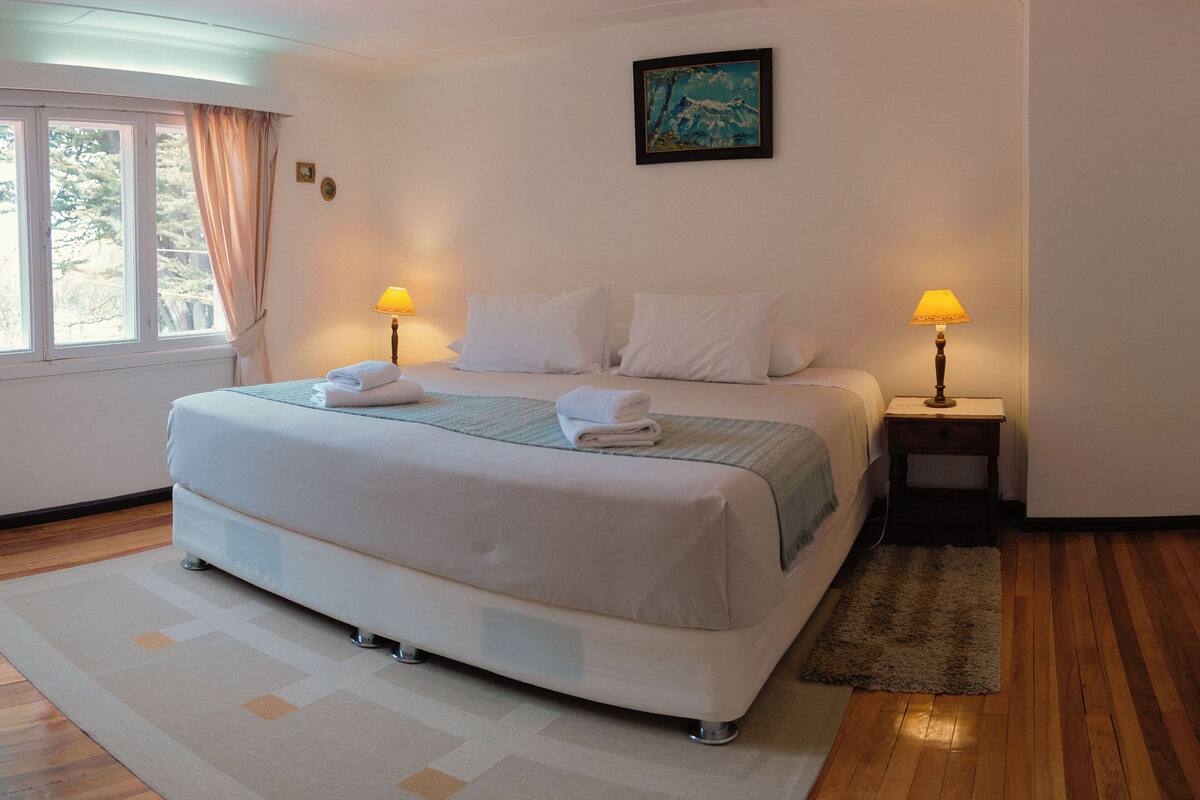 A neatly made bed with white linens, two bedside lamps, a painting on the wall, and a window with curtains.