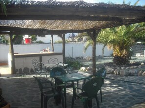 Eine Terrasse im Freien mit einem Reetdach, Steinböden und grünen Metallstühlen.